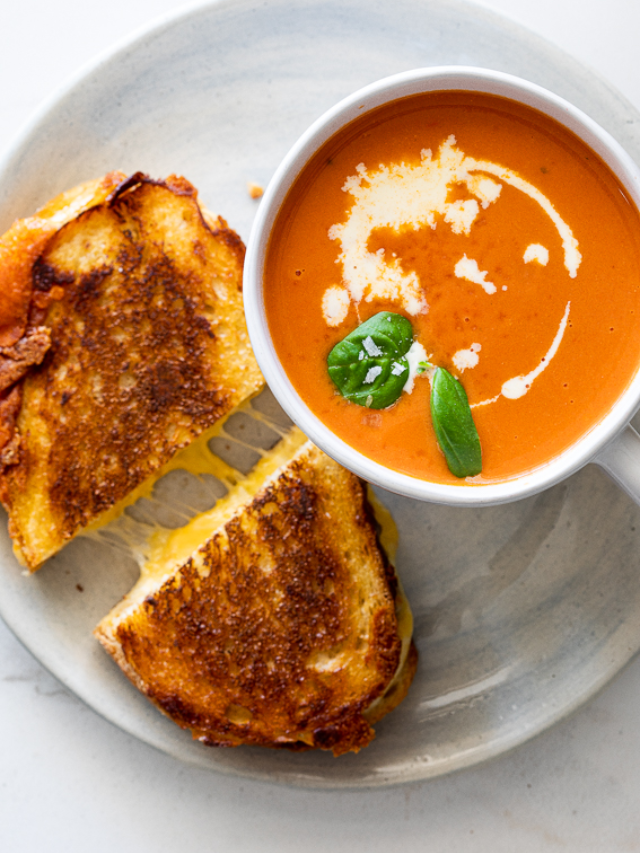 A steaming mug of tomato soup with a melted grilled cheese sandwich on the side.
