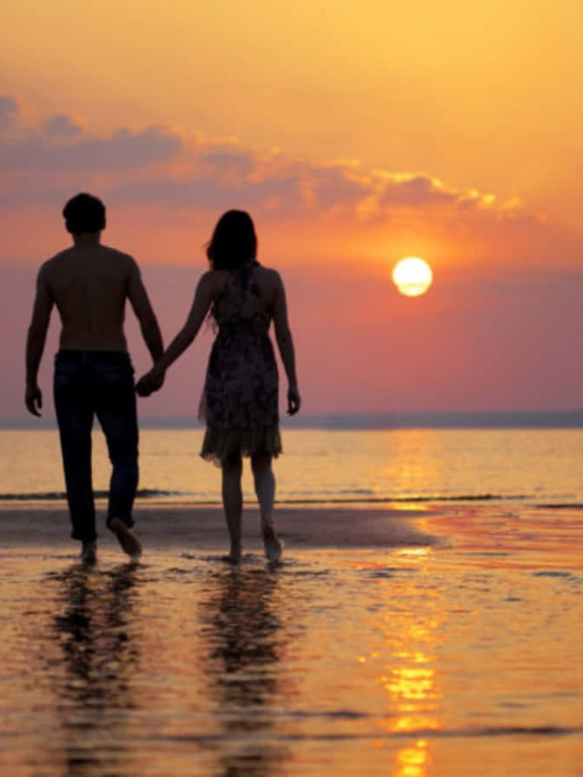 A couple walking hand-in-hand on a beach at sunset, with the silhouette of sailboats in the distance.

