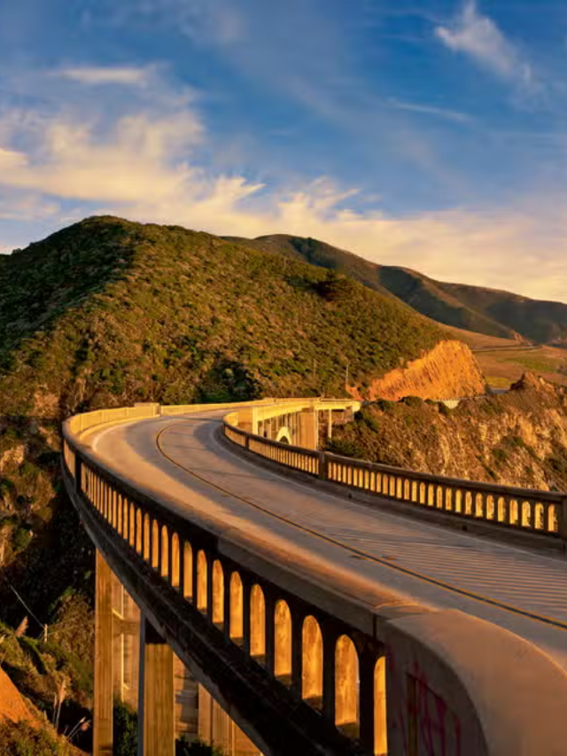 A car driving along a winding PCH road towards a sunset over the Pacific Ocean.
