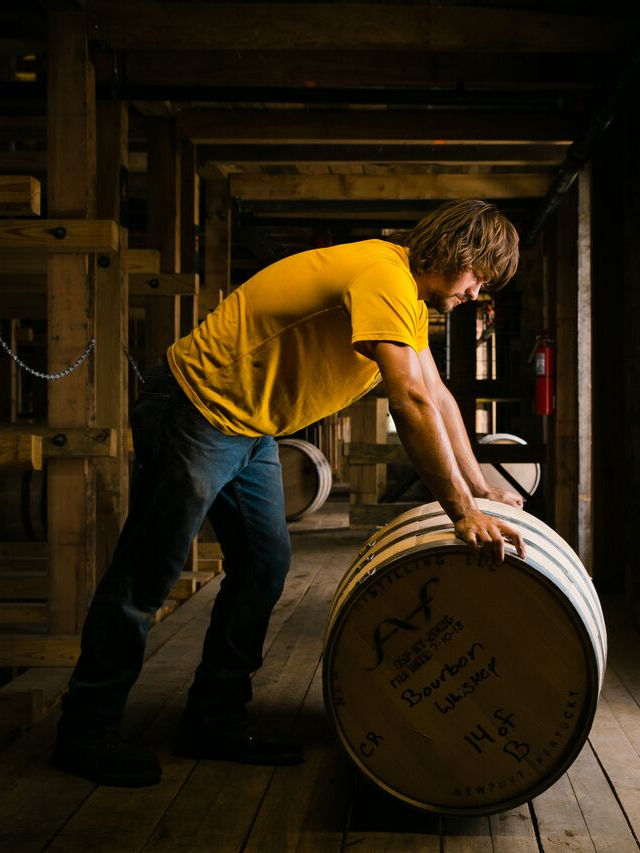 Photorealistic image of a steaming glass of bourbon next to a barrelhouse with charred oak barrels.
