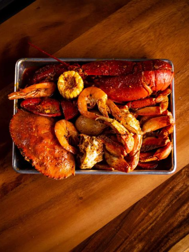 A close-up image of a plate piled high with steamed blue crabs, Virginia ham with biscuits, and a glass of red wine.
